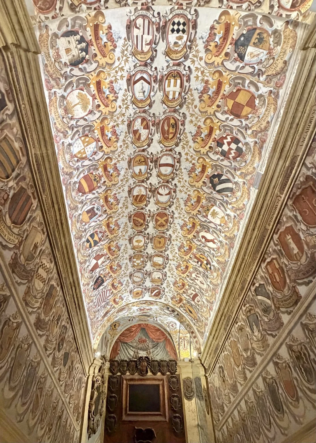 The vaulted ceiling and walls of the Archiginnasio, densely decorated with colourful coats of arms.