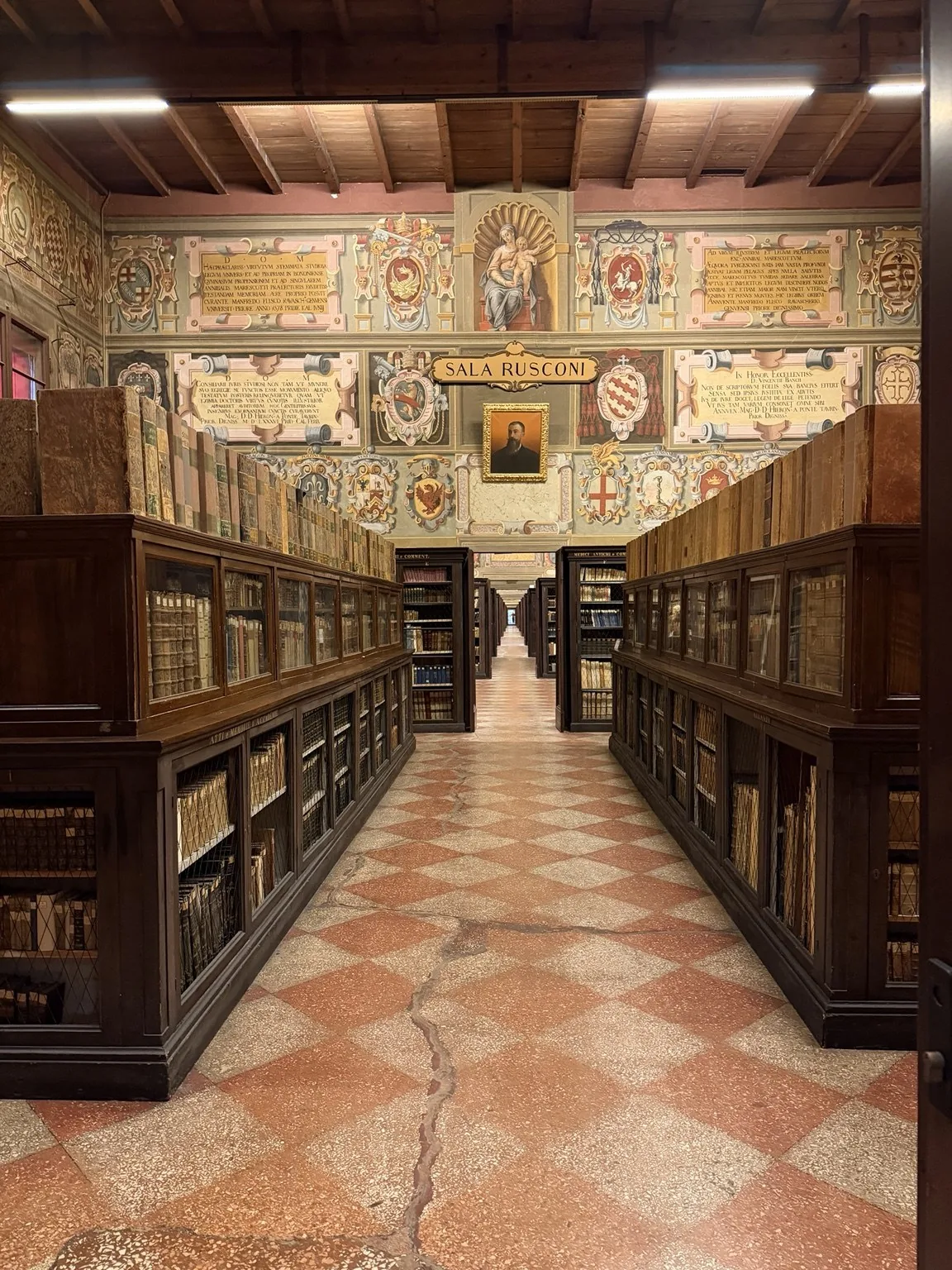 A room with shelves of antique books, a chequered floor and walls decorated with coats of arms.