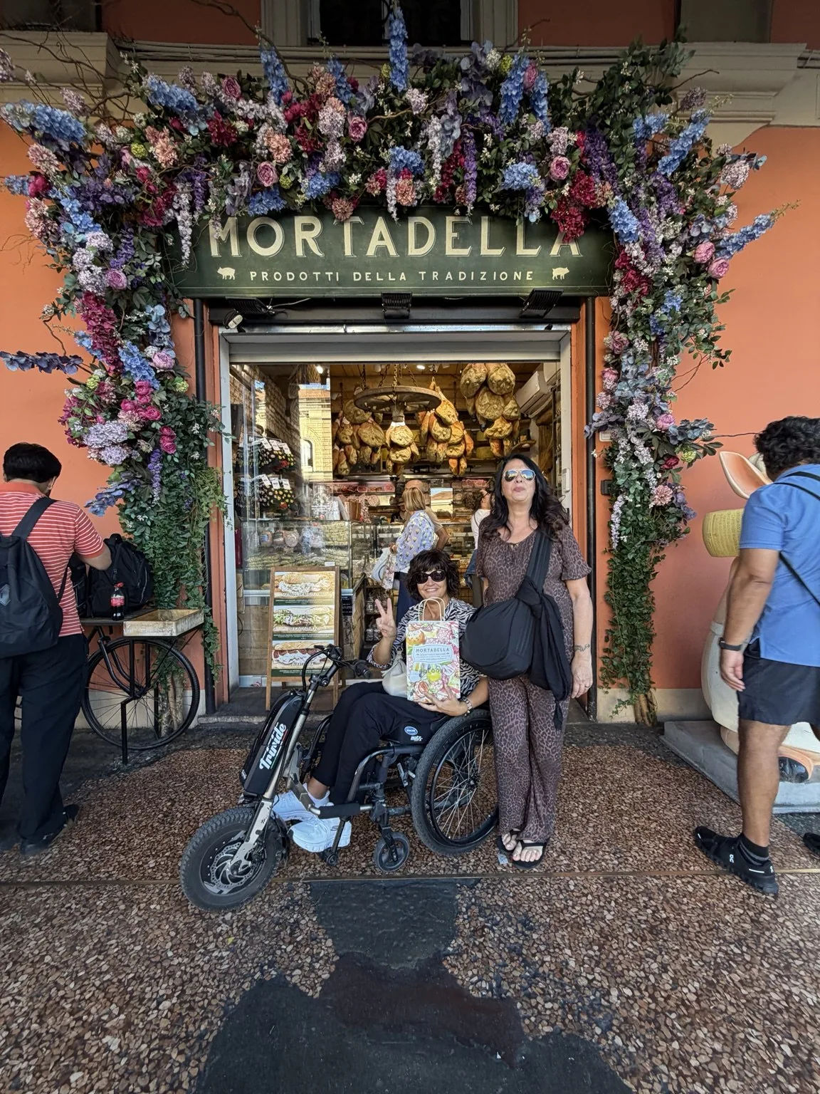 Anna on the Triride in front of a mortadella shop with a doorway framed in flowers.