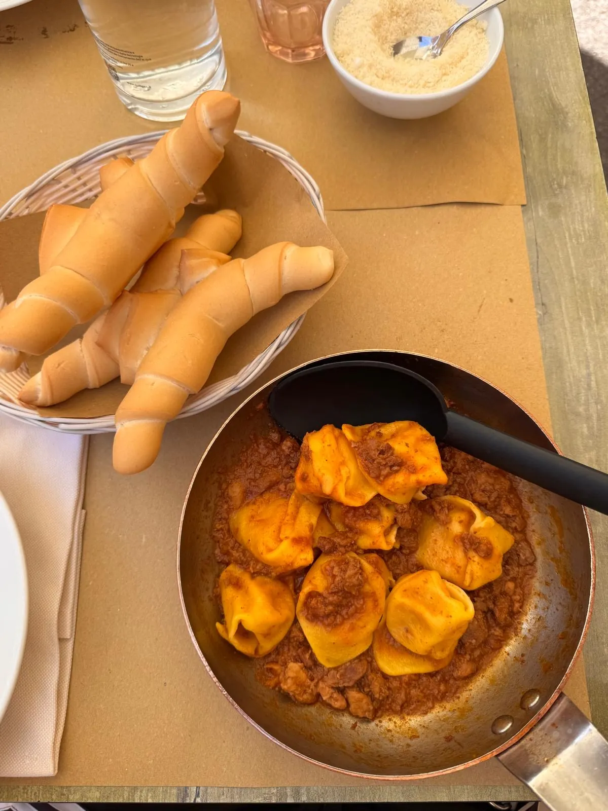 Una padella di cappellacci di zucca con il ragù, accanto a un cestino di pane ferrarese e a una ciotola di formaggio grattugiato.
