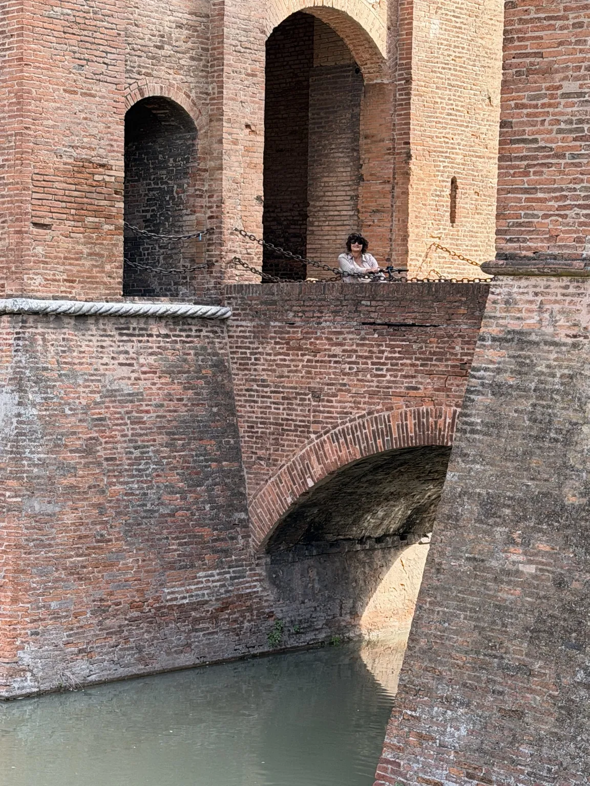 Le alte mura in mattoni del Castello Estense sopra l’acqua verde del fossato; in alto, piccola tra le arcate, Anna si affaccia da un ponte.