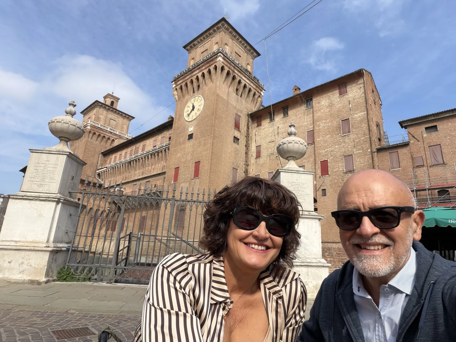 Anna e il suo accompagnatore sorridono in un selfie davanti al Castello Estense, con la torre dell’orologio e le colonne di pietra all’ingresso.