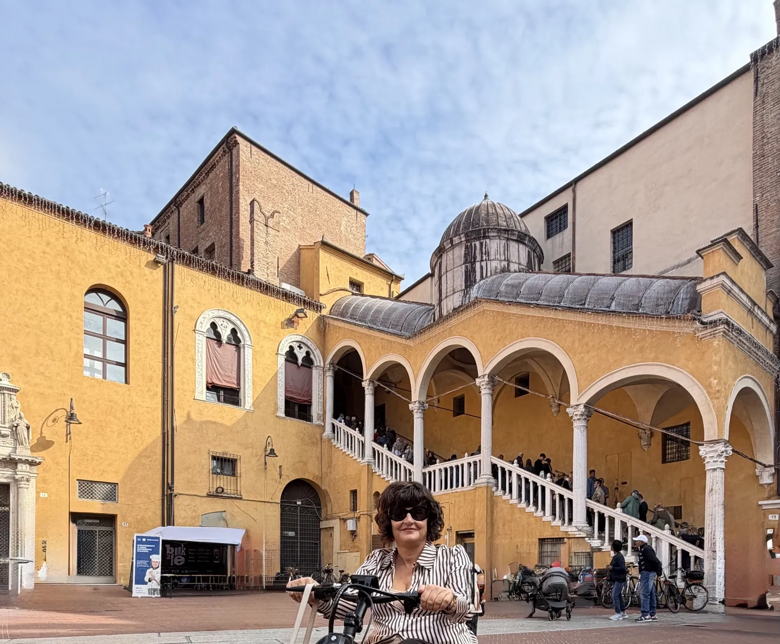 Anna sul suo Triride in Piazza Municipale, davanti al palazzo giallo con lo Scalone monumentale dalla balaustra bianca e la piccola cupola.