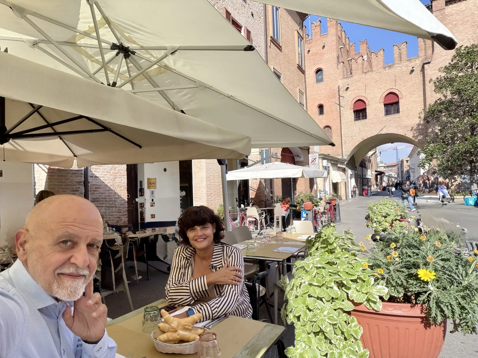 Il tavolo all’aperto sotto gli ombrelloni: l’accompagnatore di Anna in primo piano, Anna seduta di fronte e, sullo sfondo, una strada del centro con un arco merlato.