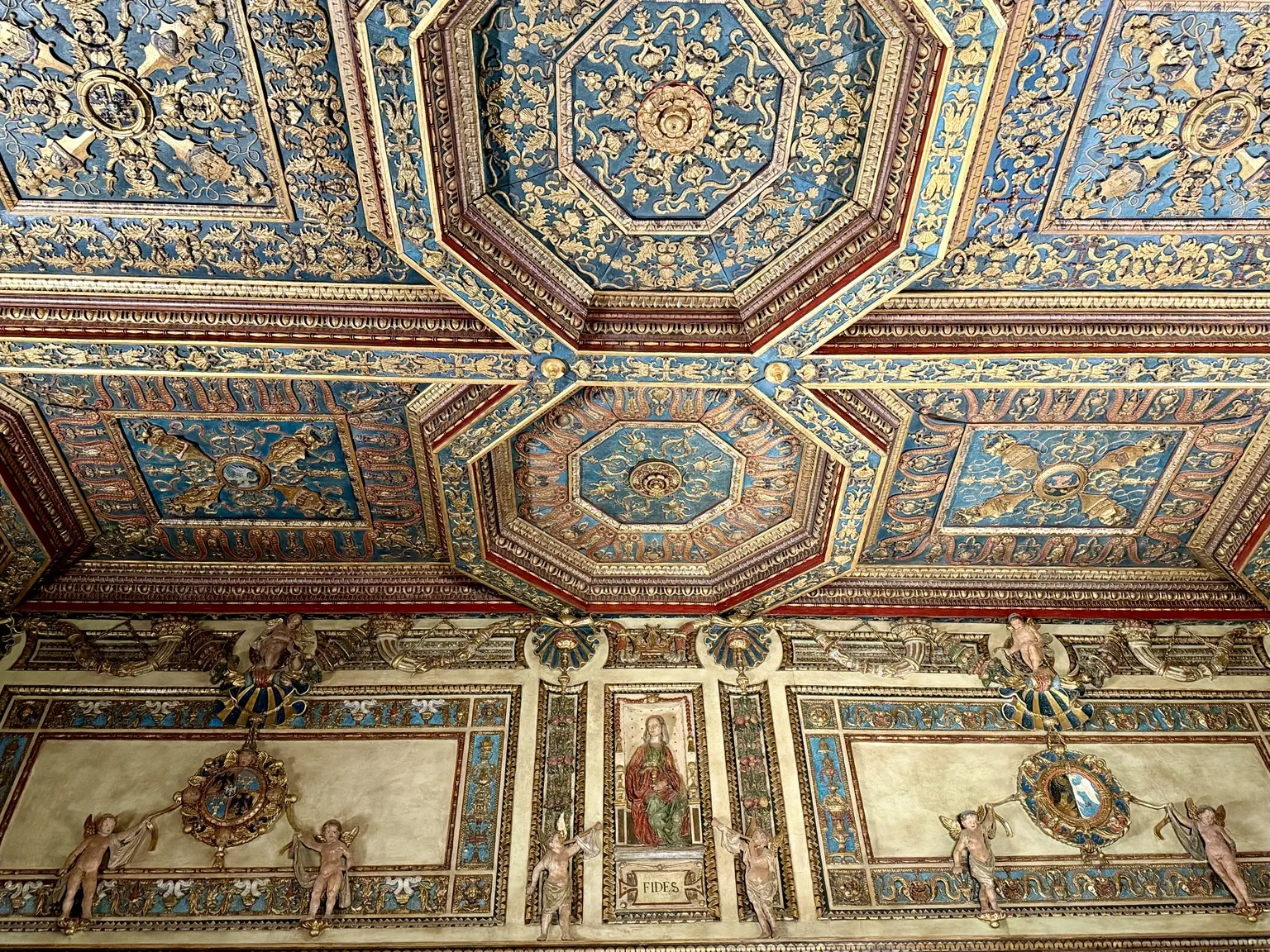 Il soffitto a cassettoni della Sala delle Virtù, con ottagoni dorati su fondo blu e rosso; sotto, la fascia alta delle pareti con stucchi, angioletti e figure.
