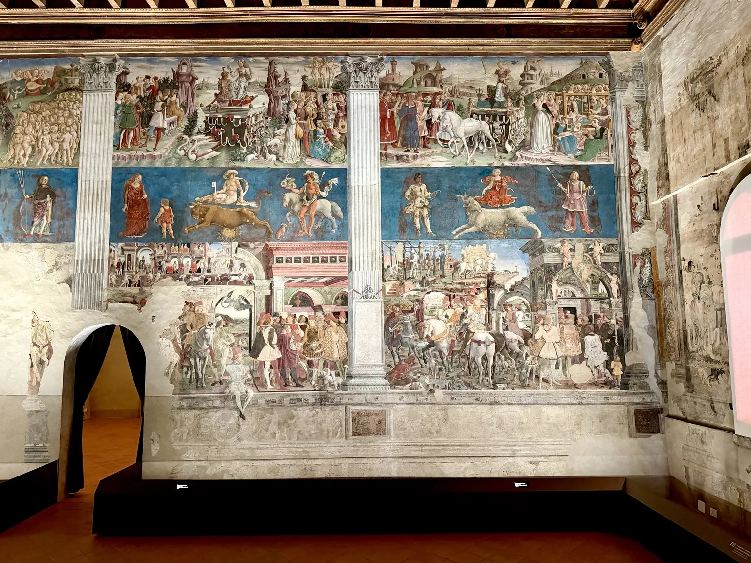 Una parete del Salone dei Mesi affrescata in tre fasce: in alto cortei e carri, al centro figure dello zodiaco su fondo blu, in basso scene di corte con cavalli e cavalieri.