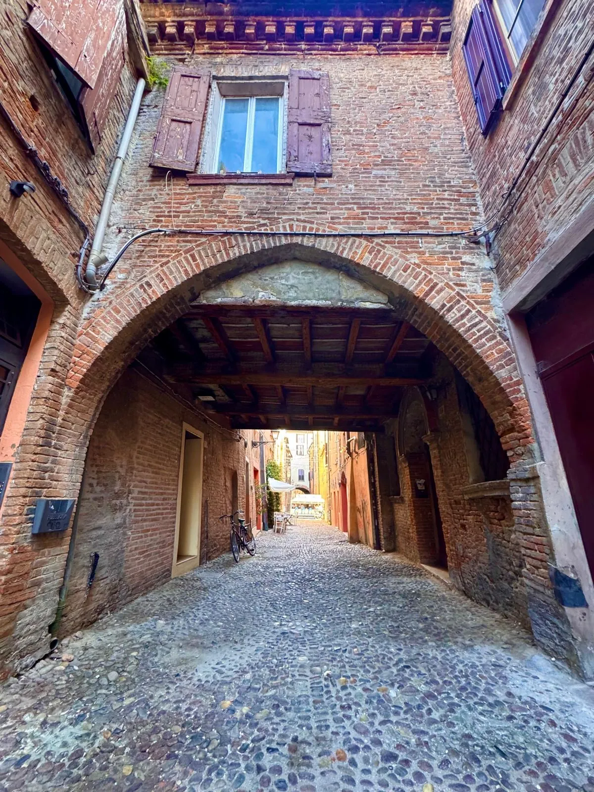 Un grande arco in mattoni con il soffitto a travi di legno sopra Via delle Volte; in alto una finestra con le persiane, sotto la strada in ciottoli.