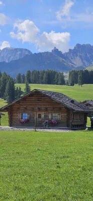 A wooden chalet on a green meadow, with Dolomite peaks in the background.