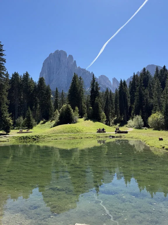 A pool of clear water surrounded by spruce trees, with the Sassolungo in the background.