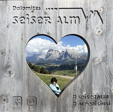 A wooden frame with a heart-shaped opening: through it you can see the Dolomites and Anna on the meadow.