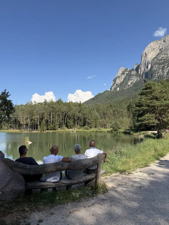 Four people seen from behind, sitting on a log bench in front of the lake, with the Sciliar in the background.