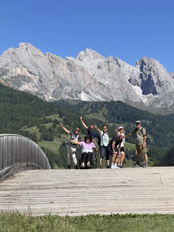 The group of friends with arms raised on a wooden boardwalk, with the Dolomites behind them; Anna is in her wheelchair in the middle.