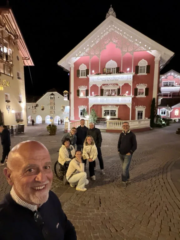 Group photo in a square in Ortisei at night, in front of an illuminated pink building; Anna is in her wheelchair in the middle.