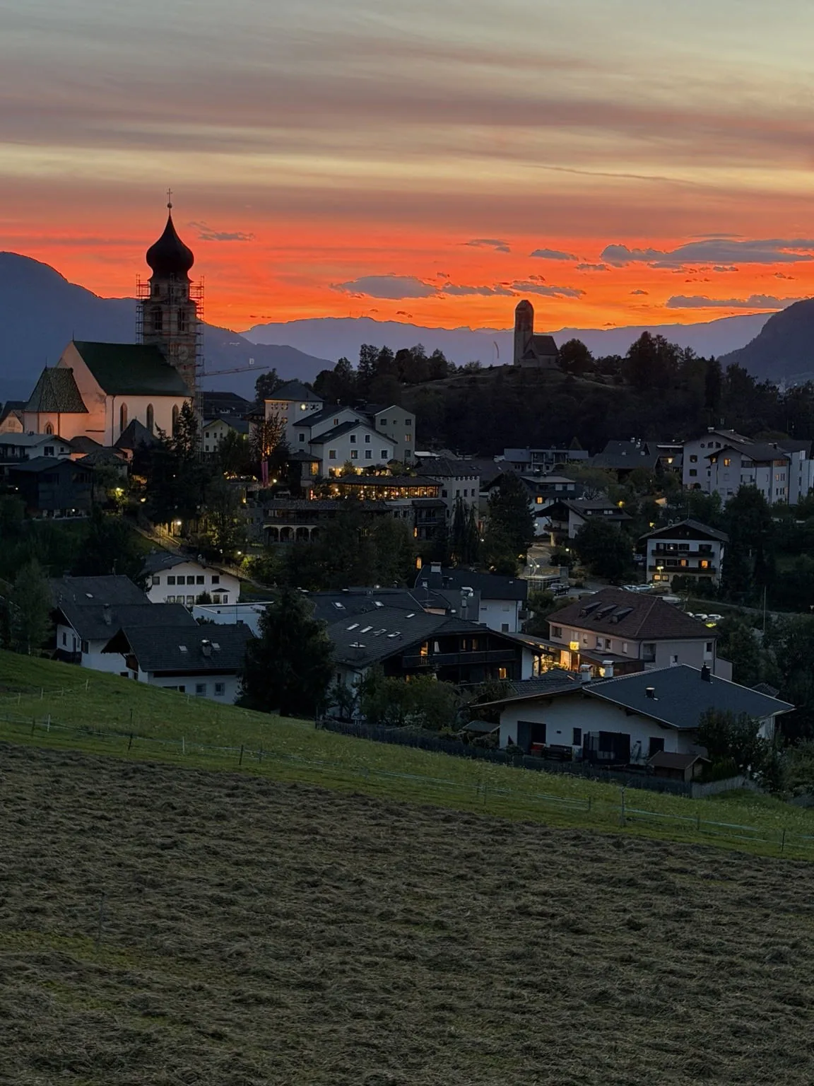 Fiè at sunset: the bell tower and houses silhouetted under a bright orange sky.