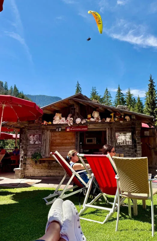 A wooden chalet, deckchairs on the lawn and a yellow paraglider in the blue sky.