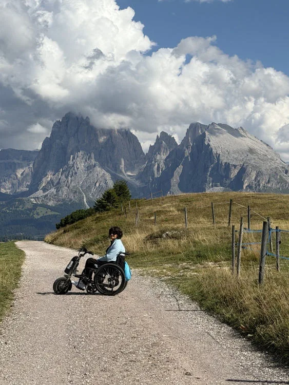 Anna con la carrozzina e il Triride su una strada tra i prati dell’Alpe di Siusi, con le Dolomiti sullo sfondo.