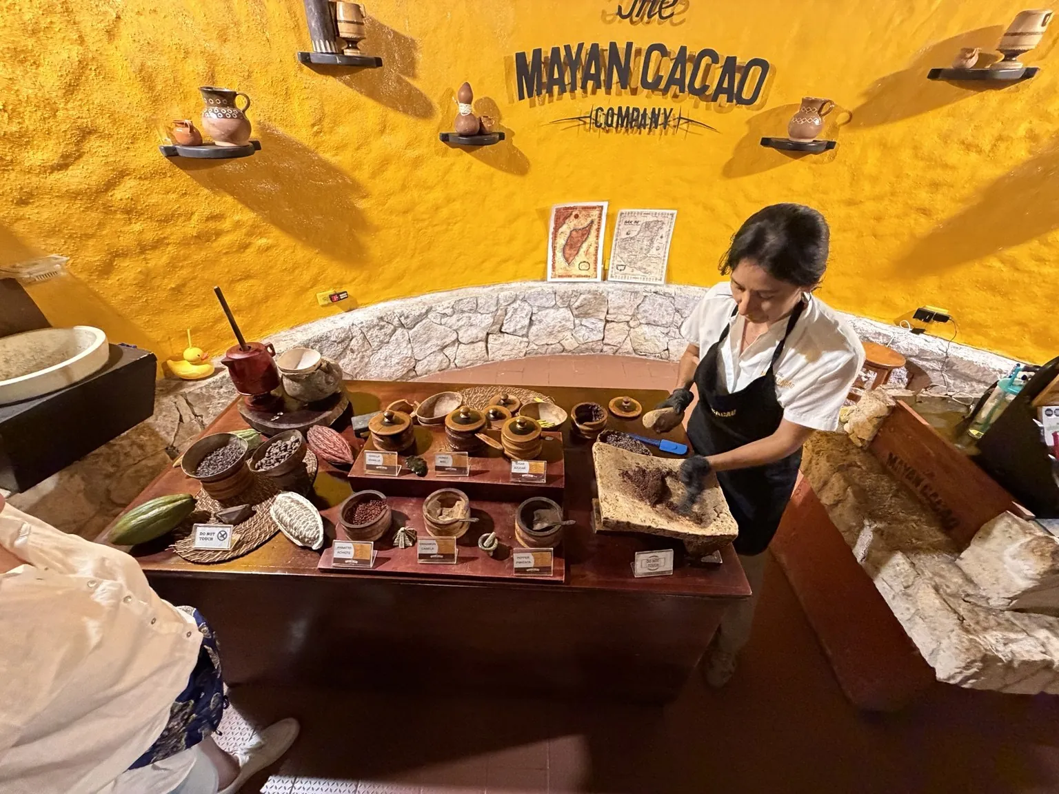 A demonstration in front of a yellow wall with the words “Mayan Cacao Company”: on a counter, bowls of beans and ingredients.
