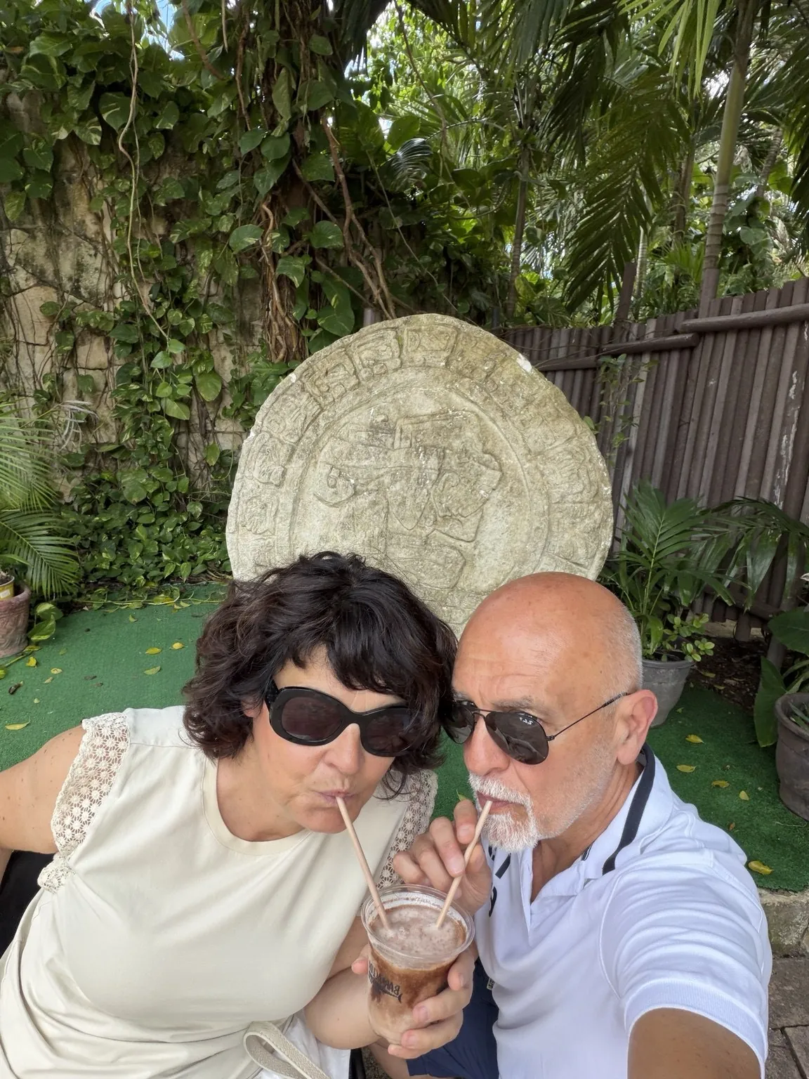 Anna and her companion drinking through two straws from the same glass, in front of a large carved stone disc.