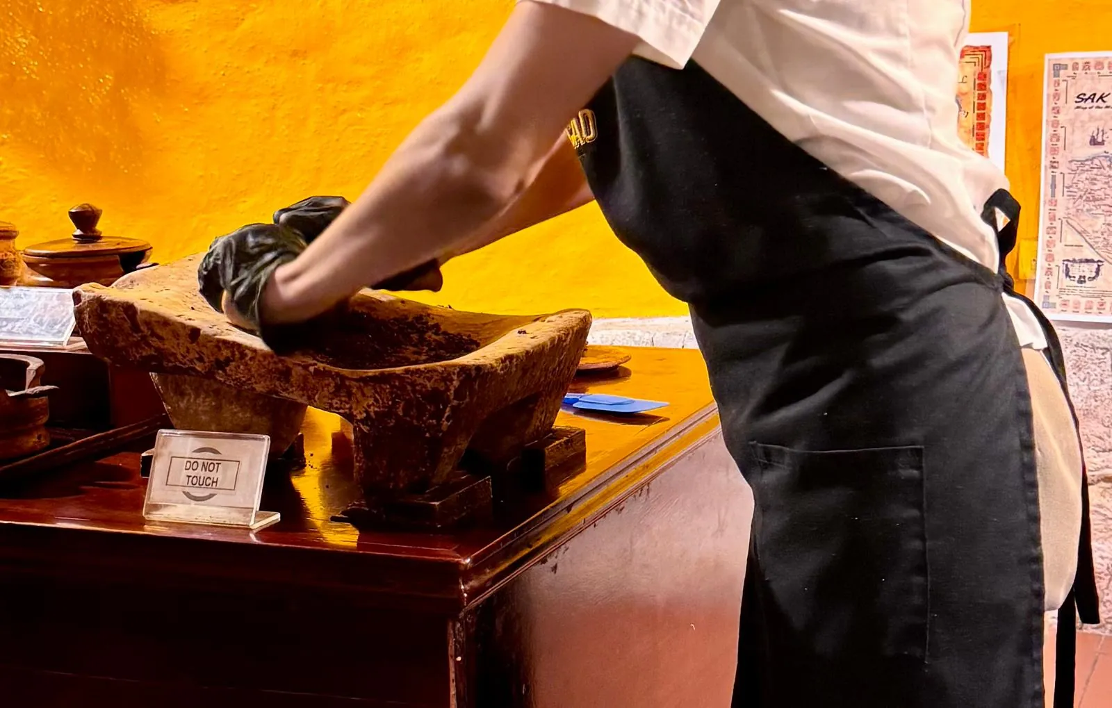 A person in a black apron and gloves grinds cacao on a sloping stone, in front of a yellow wall; on the counter, a “Do not touch” sign.