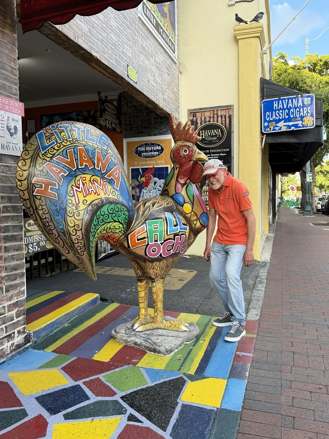 A large, brightly painted rooster statue on a mosaic pavement, in front of the signs of a cigar shop.