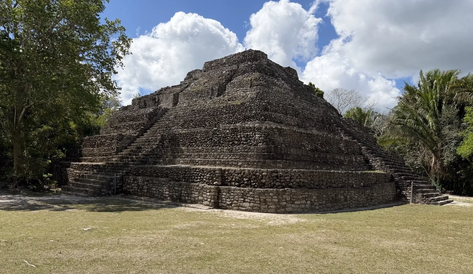 A stepped Maya pyramid in grey stone, in a clearing surrounded by trees.
