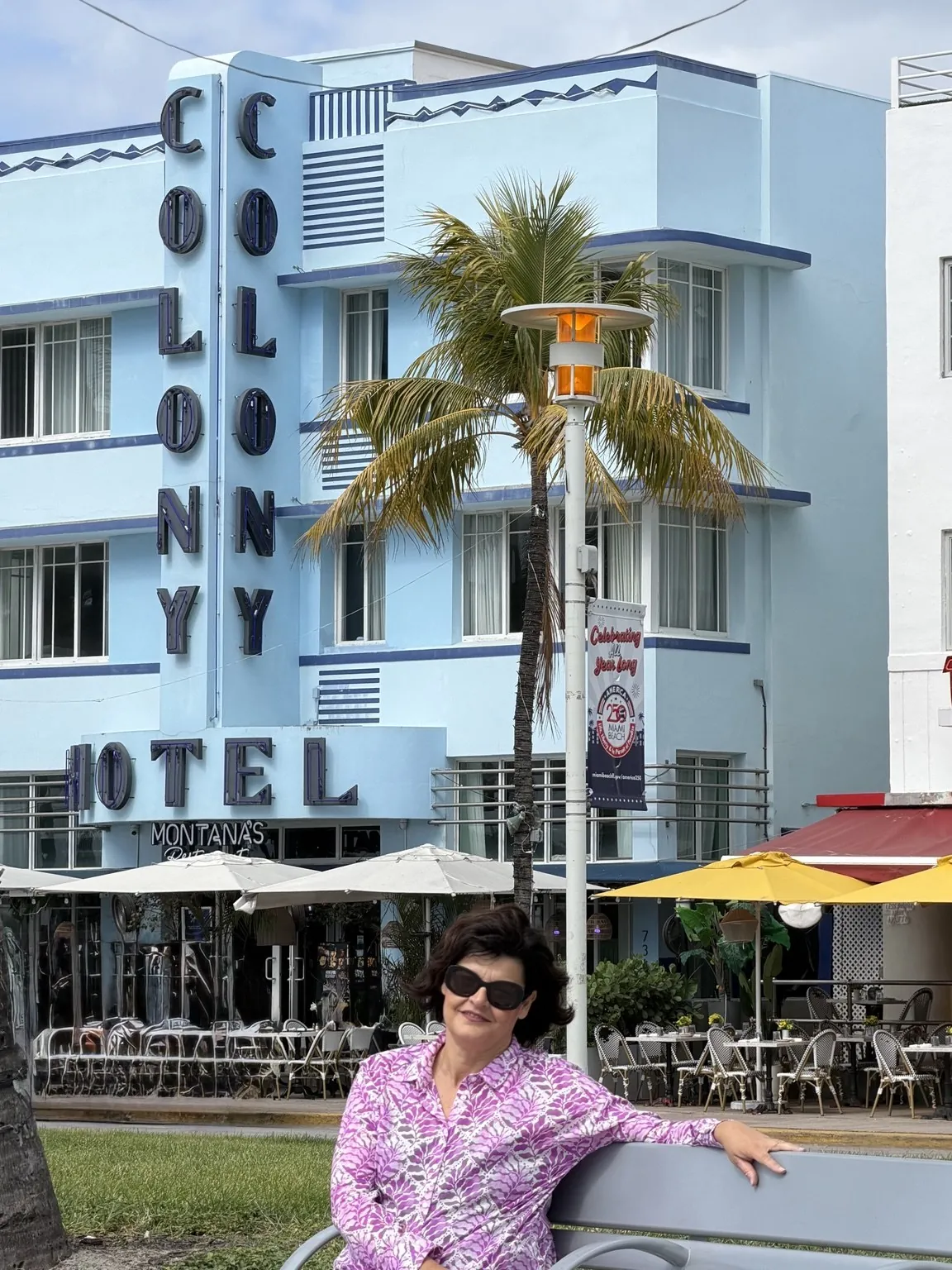 Anna sitting on a bench in front of the pale blue façade of the Colony Hotel, with its large vertical sign.