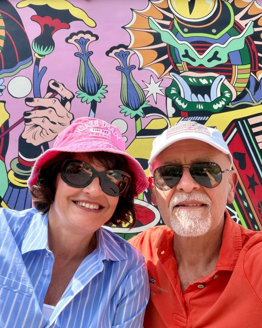 Anna, in a pink hat and sunglasses, and her companion, in a white cap and orange polo shirt, smile in a selfie in front of a large, brightly coloured mural.