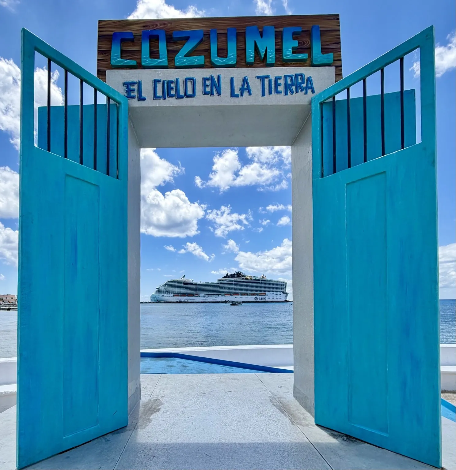A gateway with two open blue doors and the word “Cozumel”: through it you can see the sea and the ship.