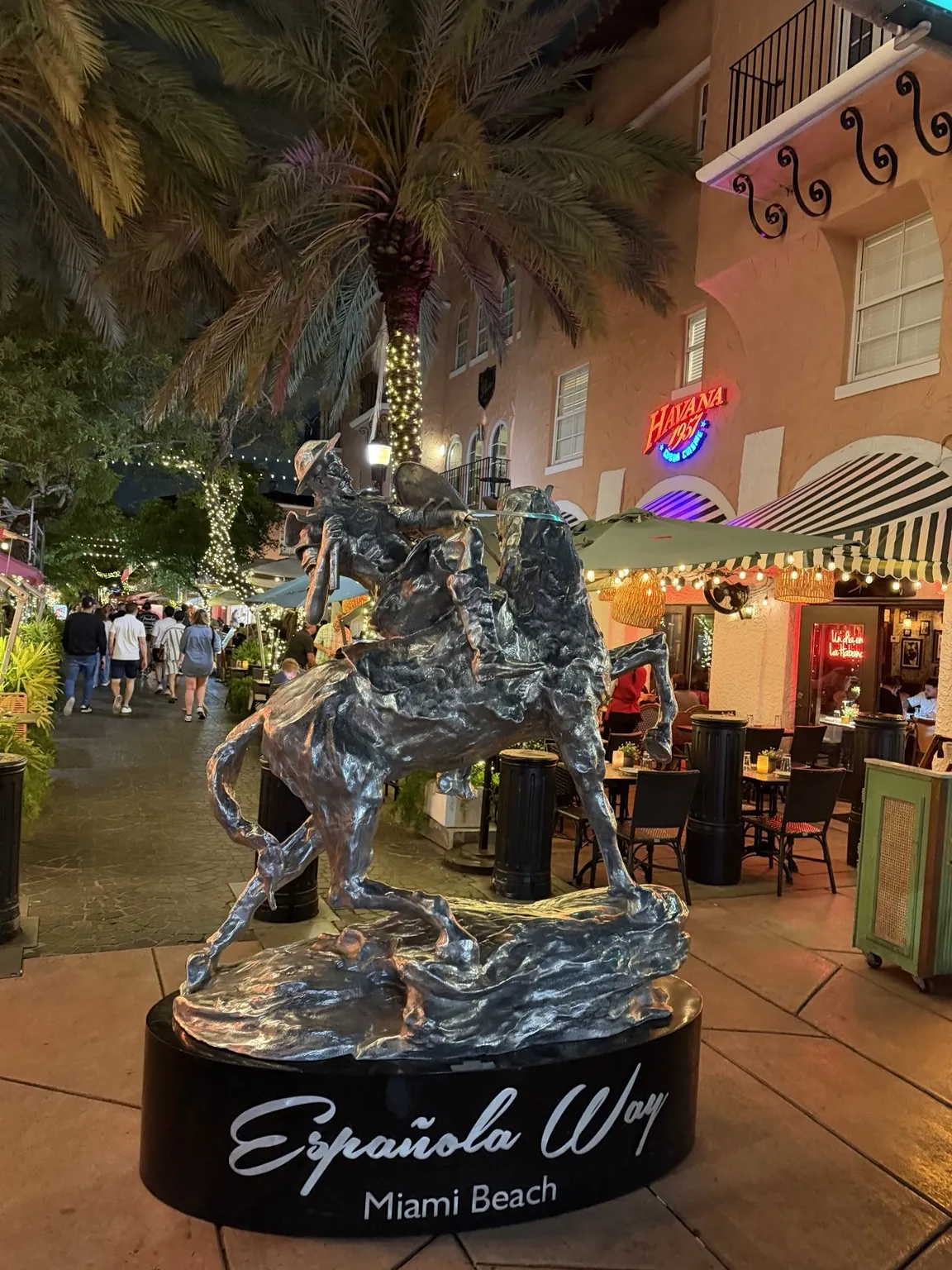 A silver sculpture with the words “Española Way, Miami Beach”, among palm trees, signs and restaurants lit up in the evening.