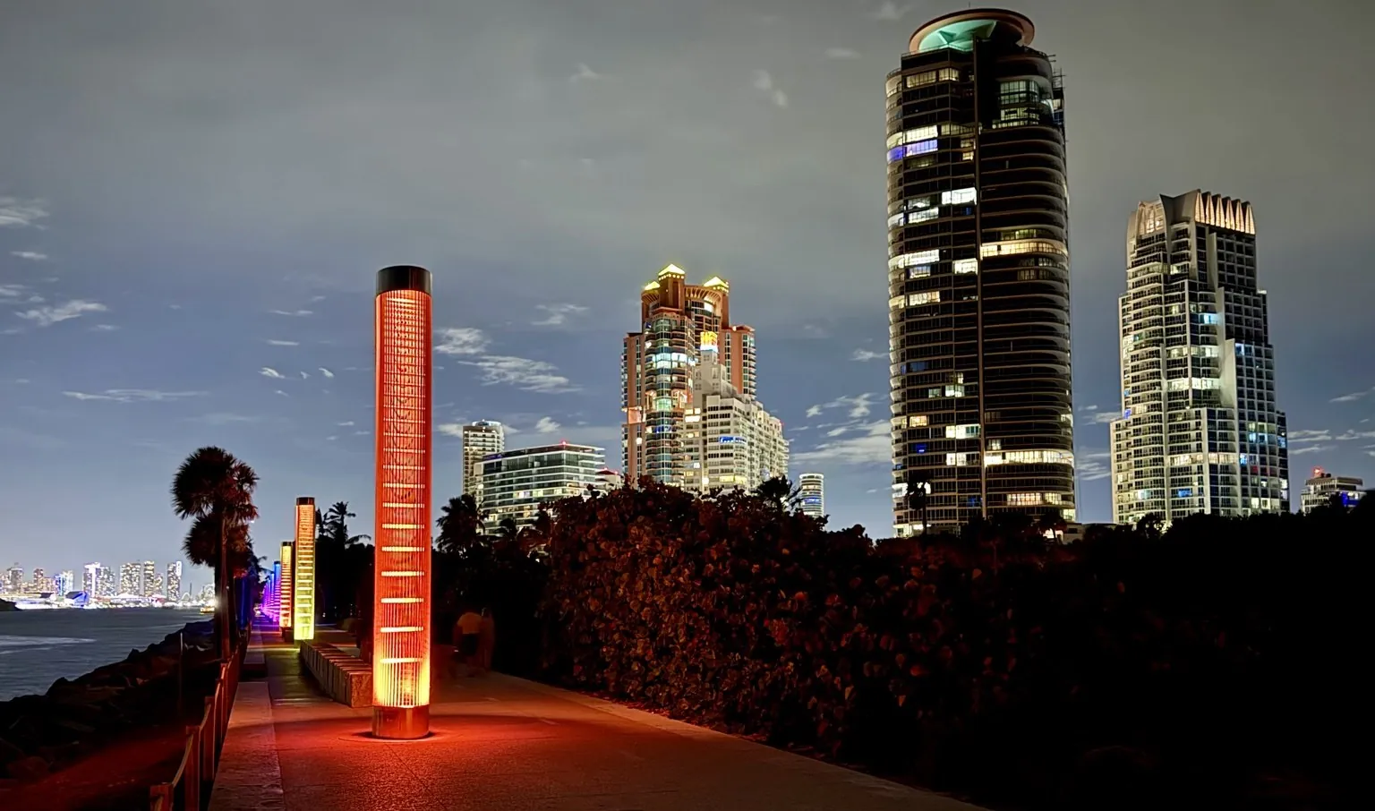 Tall skyscrapers lit up at night and a glowing orange column along the walkway.