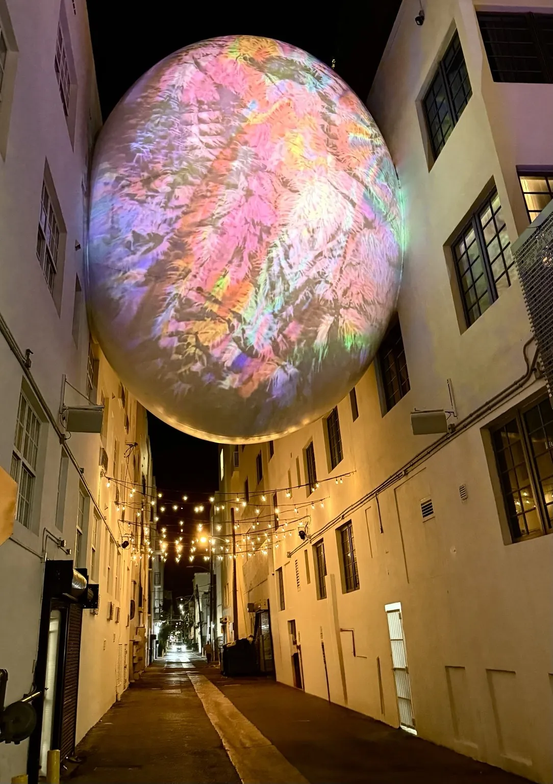 A huge glowing sphere with pink, iridescent reflections hanging between two buildings, above an alley with strings of lights.