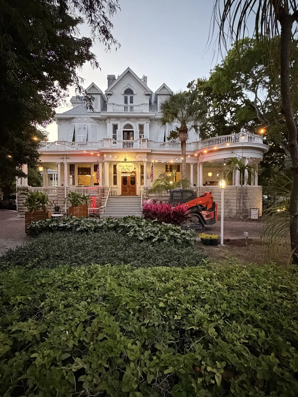 A large white house with verandas on two floors, lit up at dusk behind a garden.