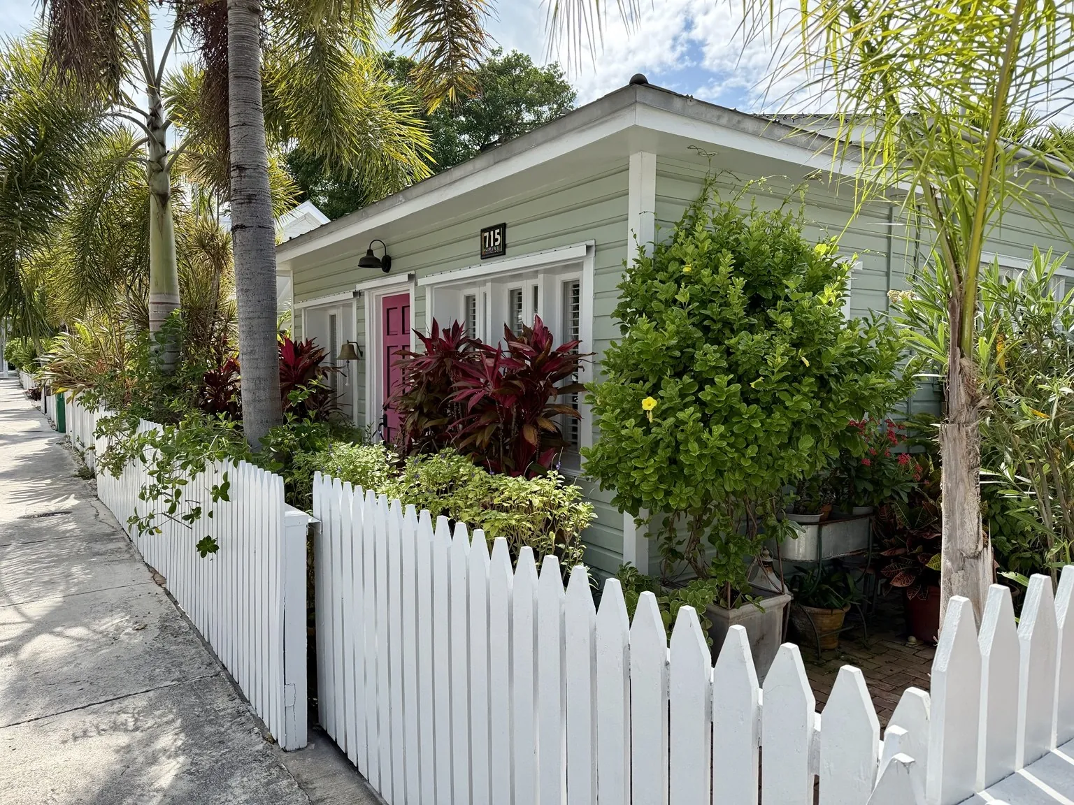 A pale green wooden house behind a white picket fence, surrounded by palm trees and tropical plants.