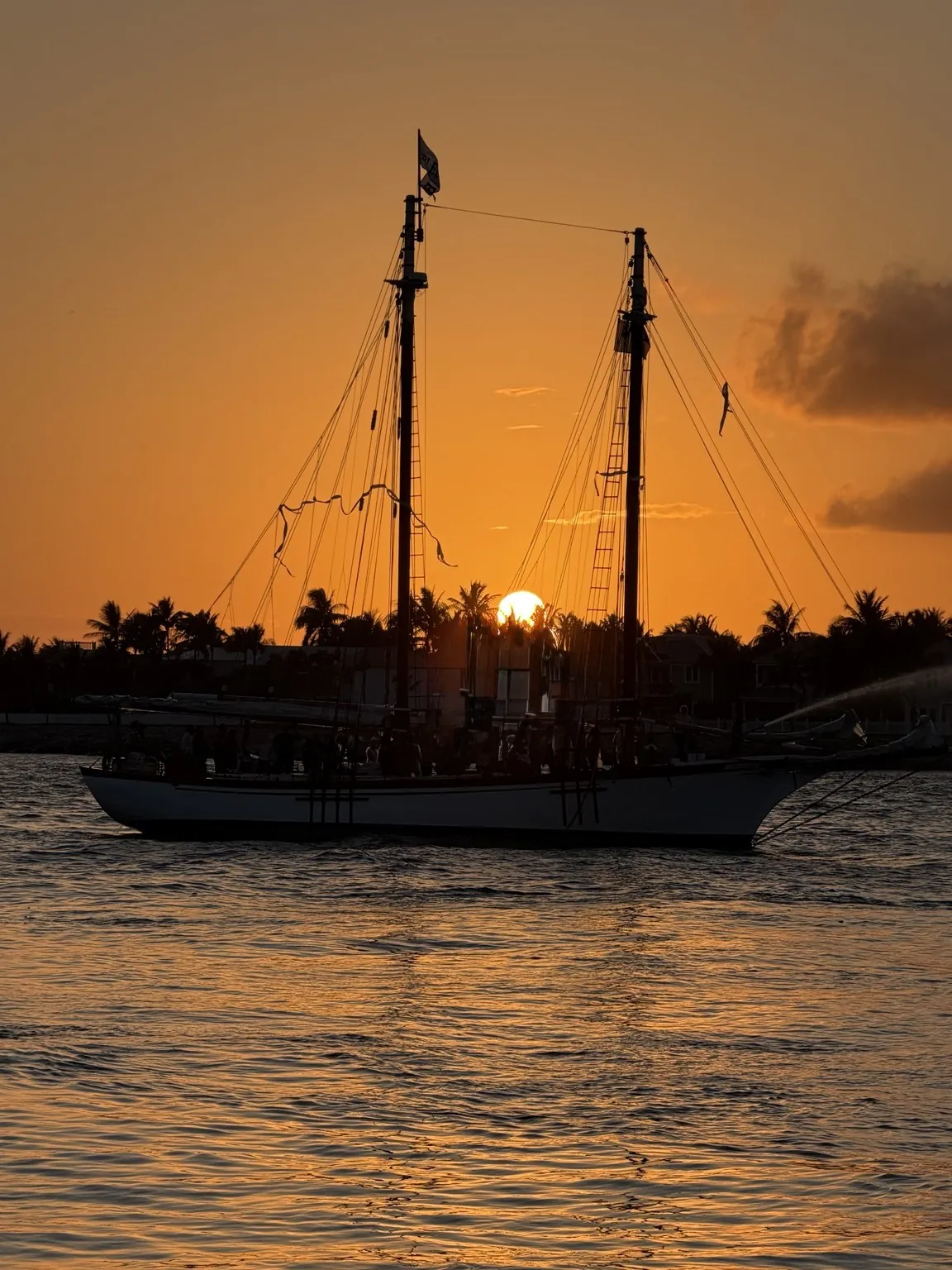 The dark silhouette of a sailing ship on the water, with the sun low and the sky orange.