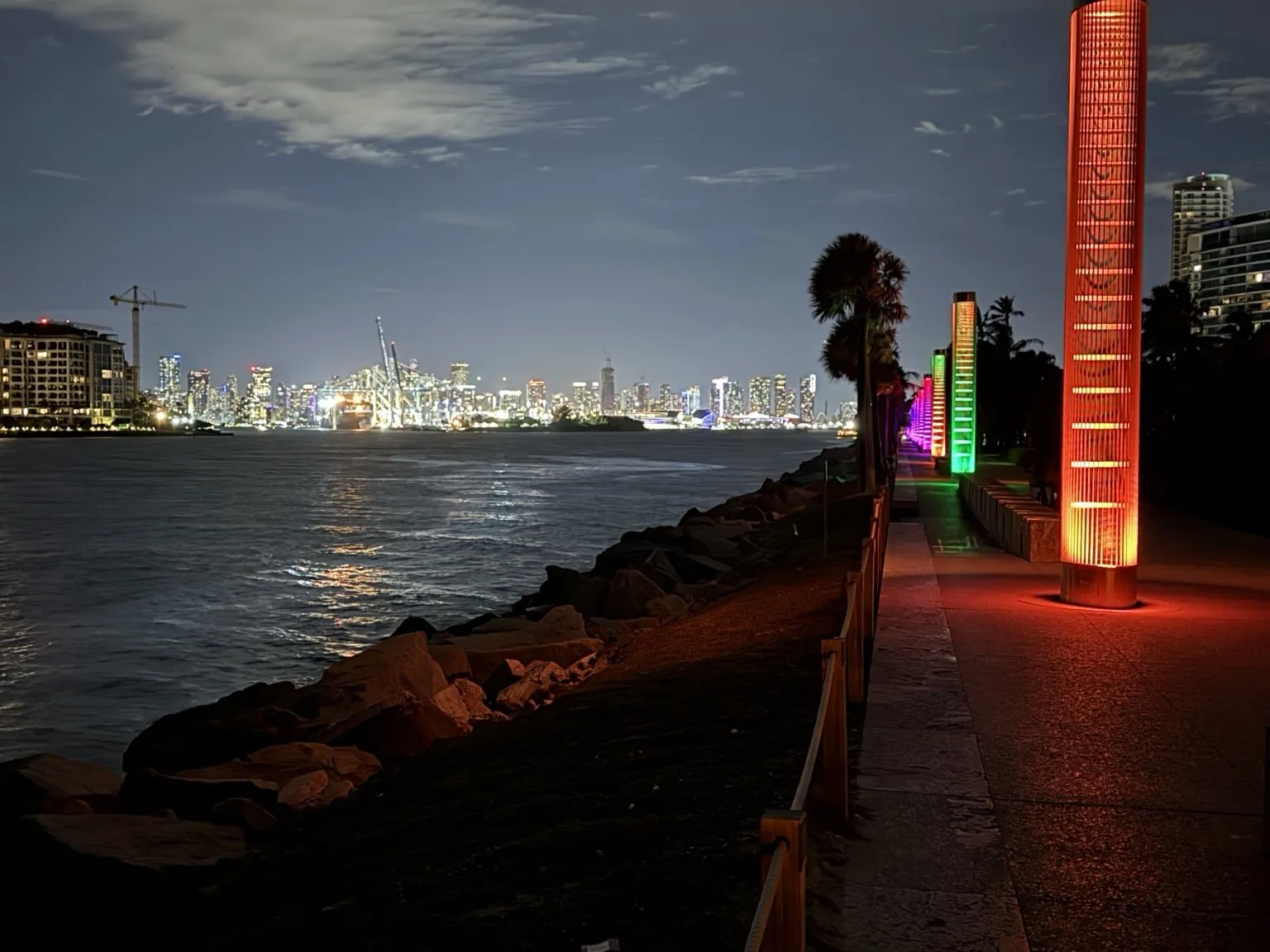 The city lights reflected on the water, seen from a walkway with colourful glowing columns.
