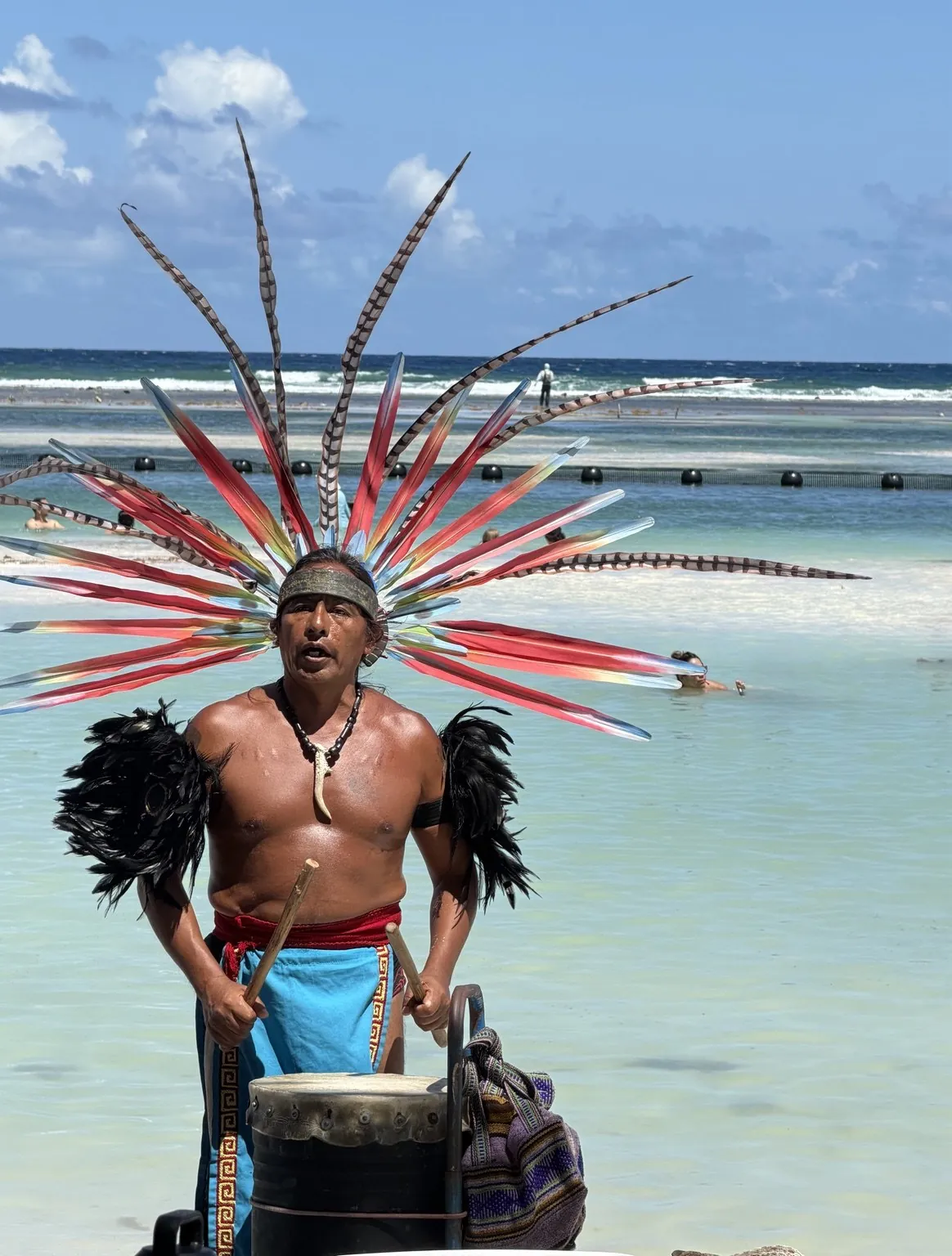 A performer in a large headdress of red and black feathers plays a drum at the water’s edge.