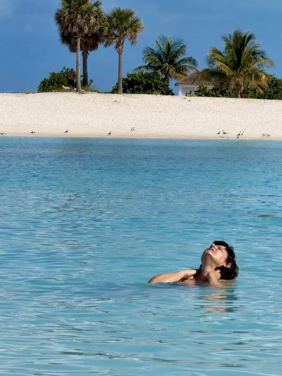 Anna in the calm turquoise water, her face turned to the sun; in the background, the white beach and palm trees.