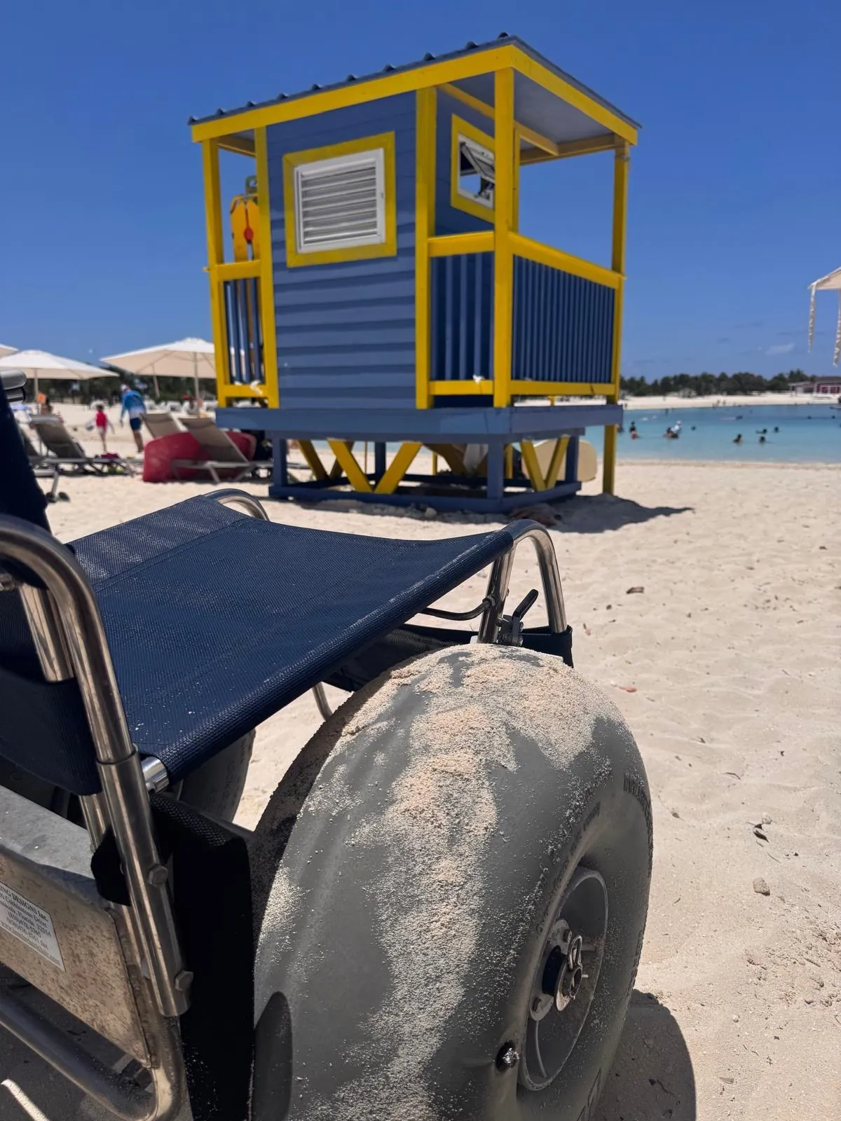 A beach wheelchair with big, wide, sand-coloured wheels, standing on the beach in front of a yellow and blue tower.