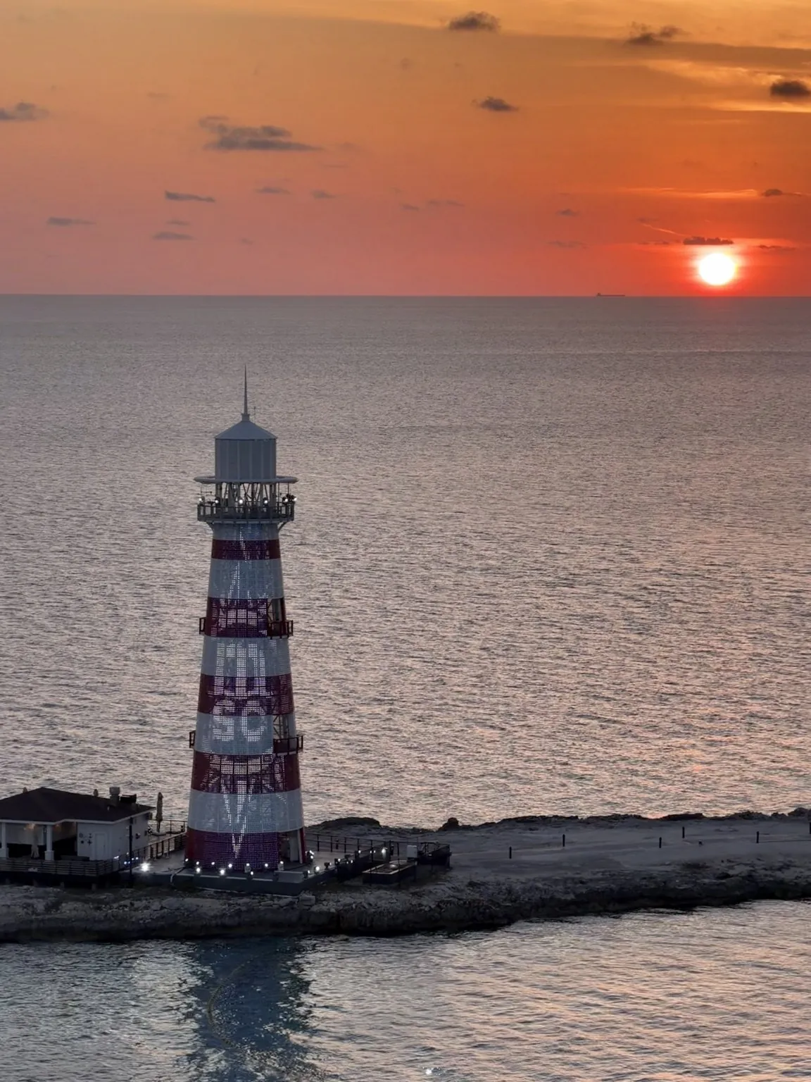 The lighthouse seen from above on the calm sea, with the orange sun low on the horizon.