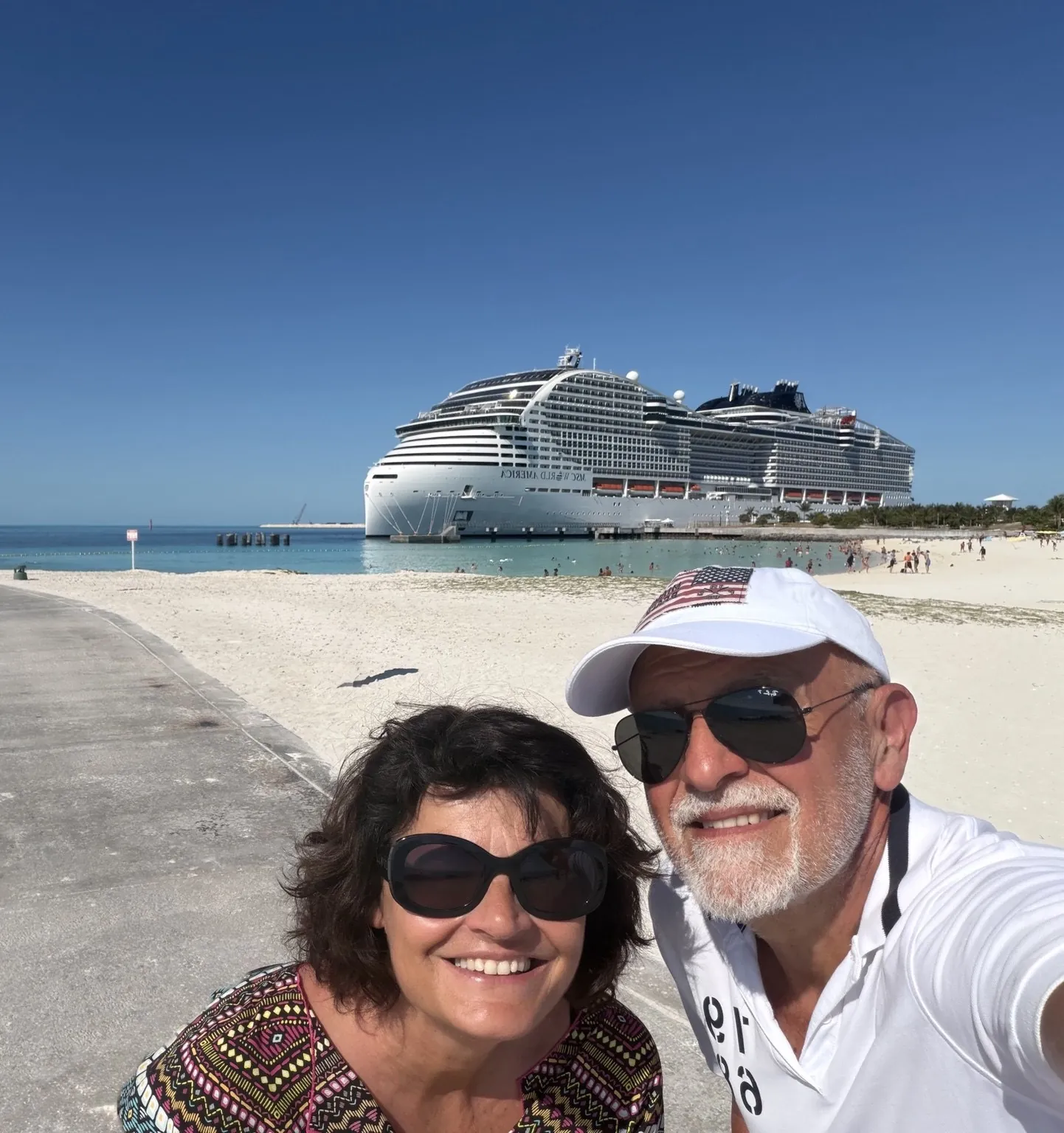 Anna and her companion smiling in a selfie on the beach; behind them, the cruise ship.