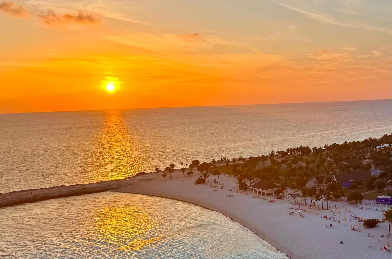 The sun setting over the sea and a strip of white beach with palm trees, lit by golden reflections.