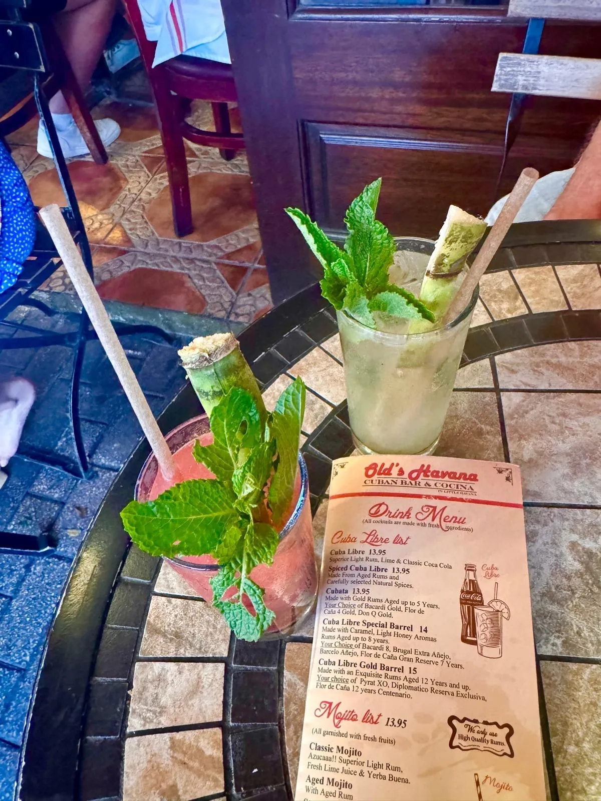 Two cocktails with mint leaves and straws on a small table, next to the bar’s drinks menu.