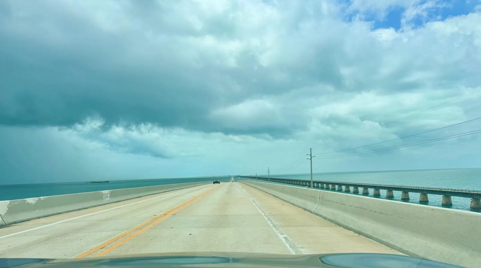 The road seen from the car: a long, straight bridge with turquoise sea on both sides, under a cloudy sky.