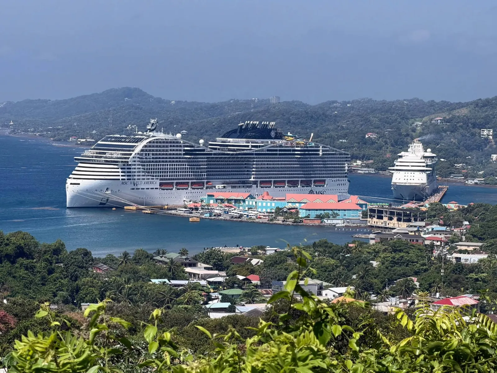 The big white MSC World America docked at the port of Roatán, seen from above through the vegetation; beside it, a second, smaller ship and, in the background, green hills.