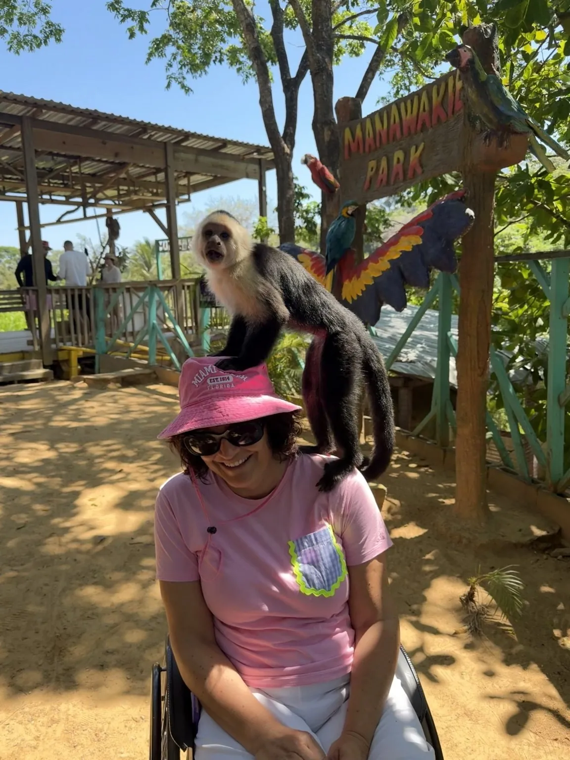 Anna, sitting in her wheelchair, laughs as a little black and white monkey climbs onto her shoulders, in front of the wooden “Manawakie Park” sign.