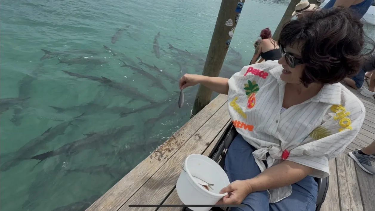 Anna leans out from the pier with a small fish in her hand; lots of big tarpon swim in the green water.