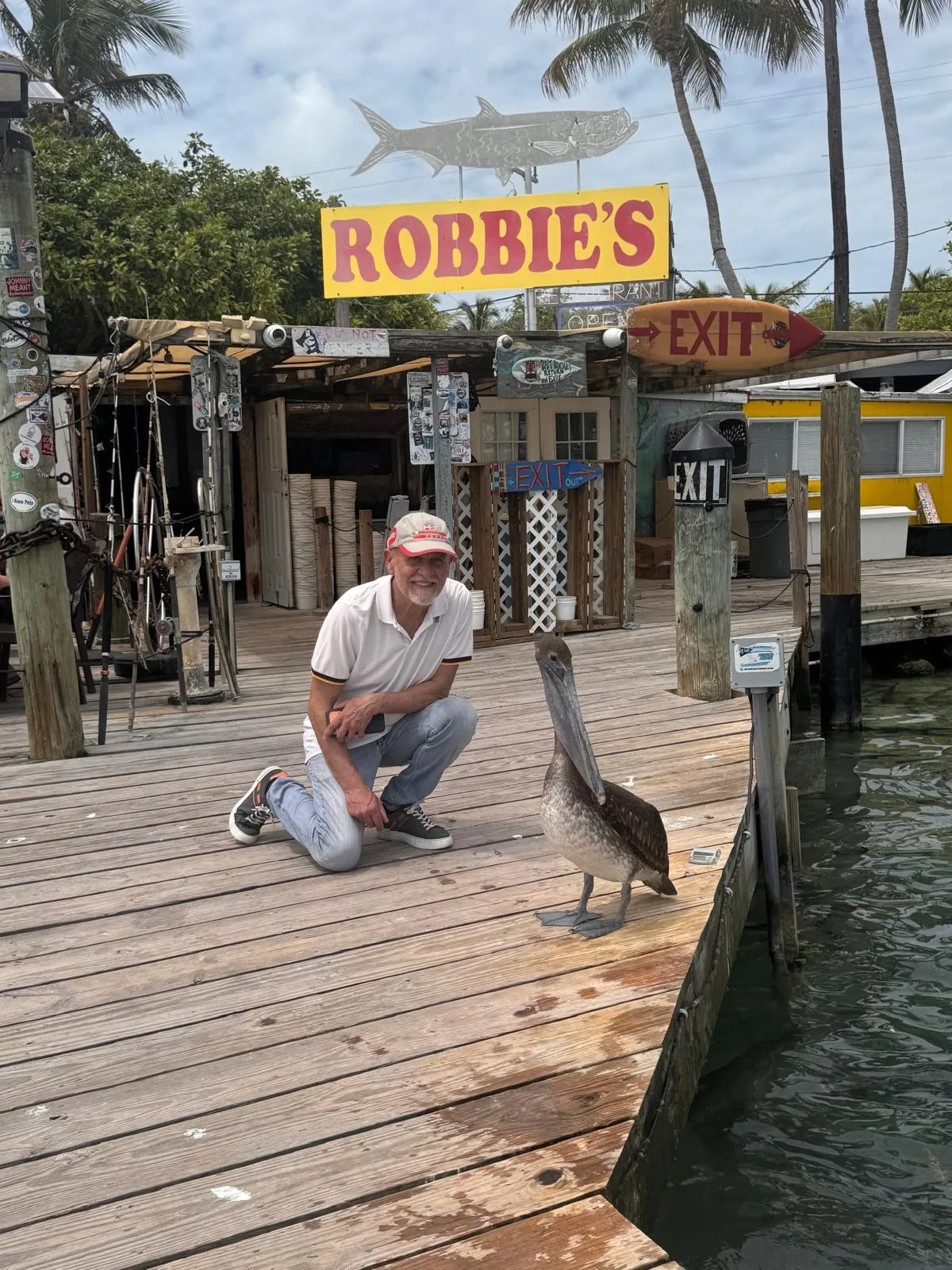 Anna’s companion crouching beside a pelican on the wooden pier, under the “Robbie’s” sign.