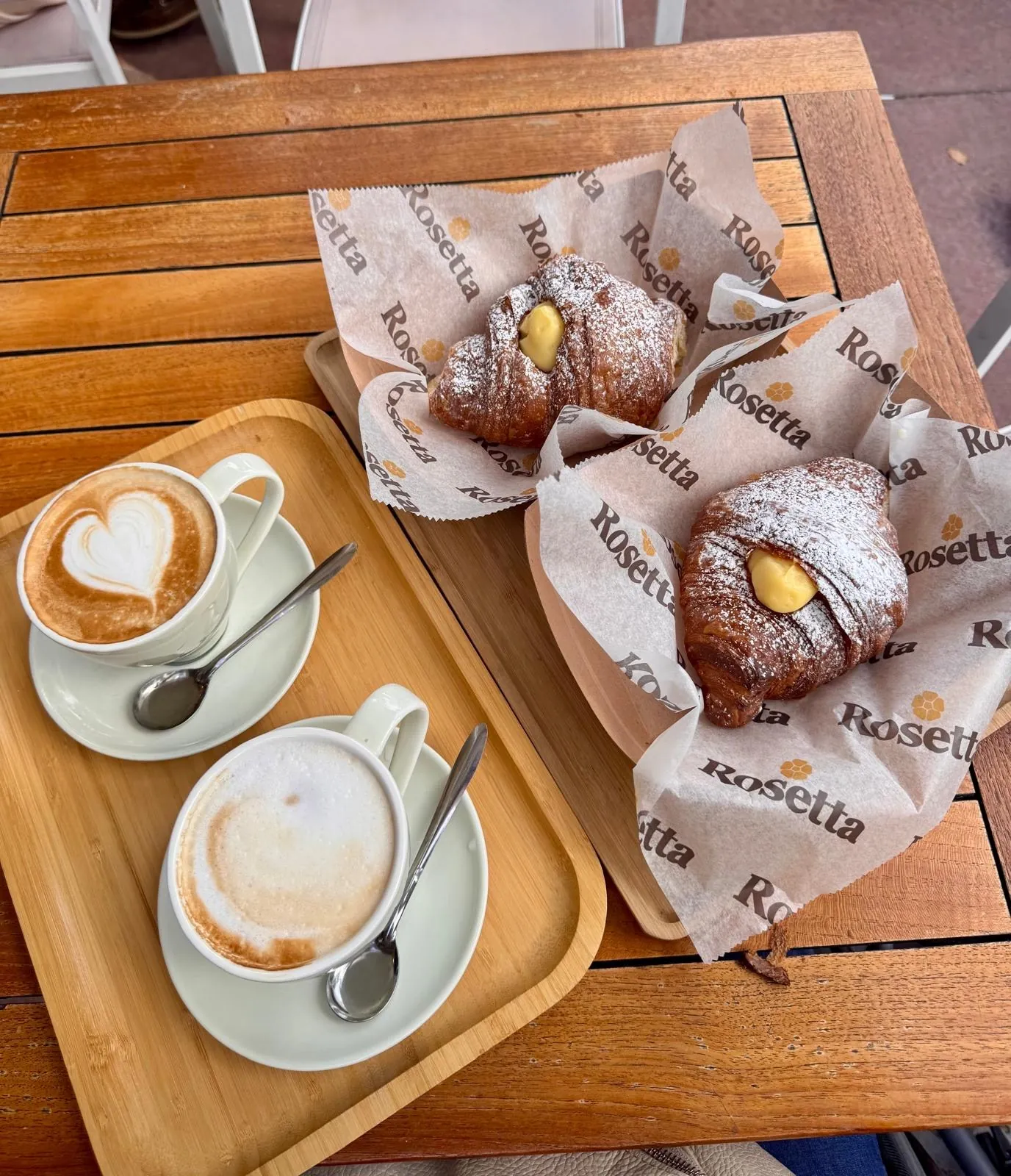 Two cappuccinos and two large filled croissants dusted with sugar, on a wooden tray.