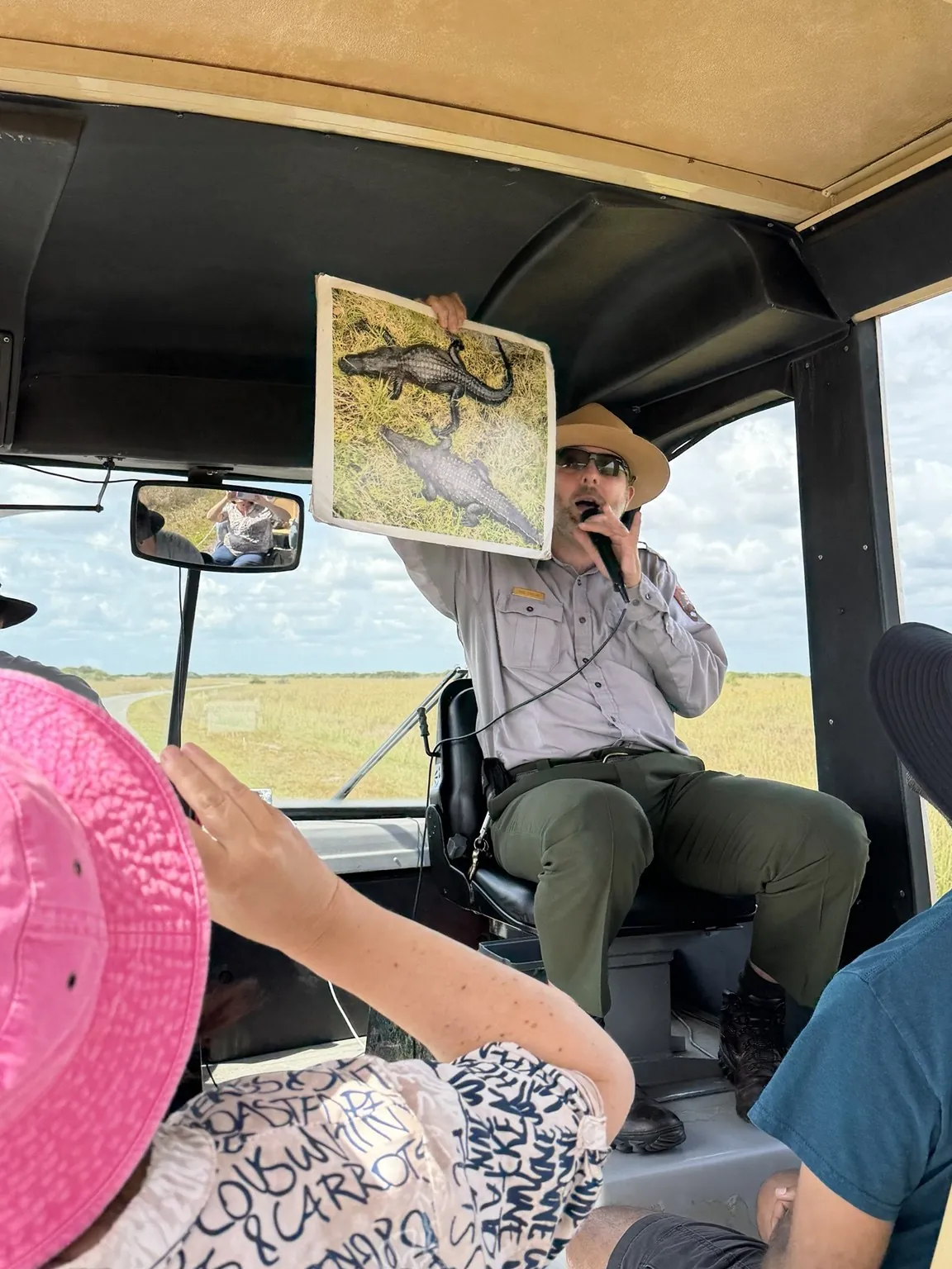 The guide, holding a microphone, shows the tram passengers an illustration of an alligator.
