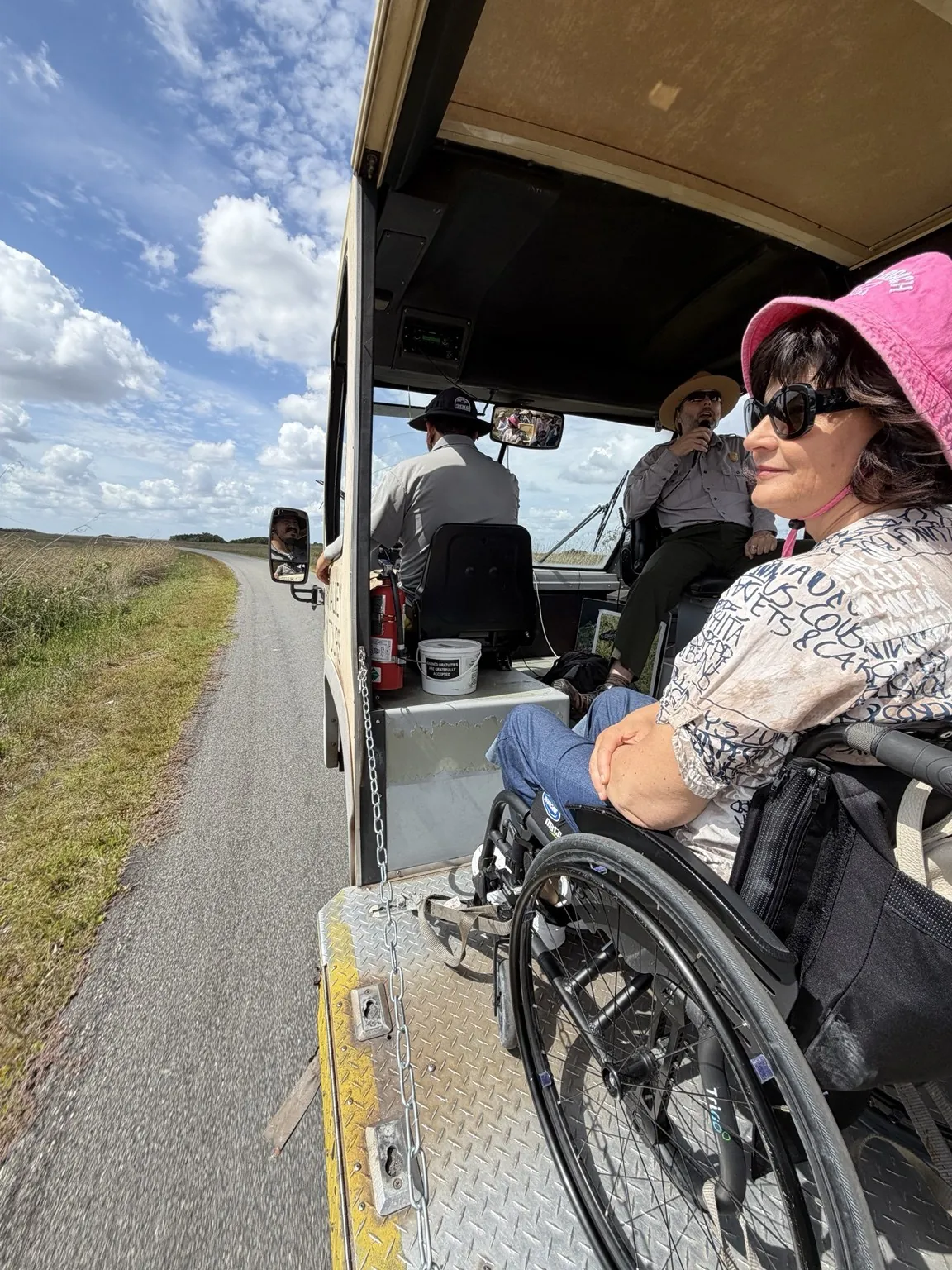 Anna, in a pink hat, sitting in her wheelchair aboard the open-sided tram; the road and the prairie roll past beside her.